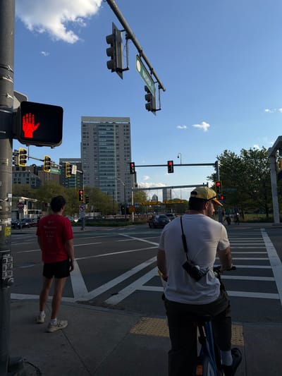 The image captures a city scene in Boston, Massachusetts, United States of America. It depicts two individuals waiting at an intersection controlled by traffic lights. One person, wearing a white t-shirt, yellow hat, and dark trousers, is on a bicycle. A black camera is slung around his neck. The second person, wearing a red t-shirt and black shorts, is standing on the sidewalk. All traffic lights visible in the frame are displaying red signals. Street signage indicates the intersection of Nashua Street and Storrow Drive. Several buildings are visible in the background, with one being a large, multi-story office building. Cars are visible on the street, and the sky is clear with a few scattered clouds. The image is rotated, with the sky towards the left.