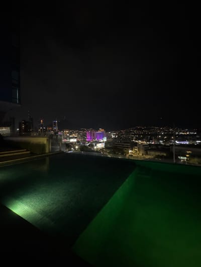 This detailed image depicts a nighttime urban landscape observed from a high vantage point, likely a rooftop. The foreground features an infinity-edge swimming pool with dark-colored tiling, illuminated from the lower left by a bright green light source, casting a distinct striped pattern across the water's surface. An adjacent dark green surface, possibly a deck or secondary pool area, is visible to the right. On the far left, a partial view of a dark building with a few lit windows is present, suggesting the structure from which the photograph was captured.
The midground reveals a sprawling cityscape with numerous buildings of varying architectural styles and heights. Many structures are illuminated, with some displaying distinct colors, notably a prominent building to the mid-left showcasing vibrant purple lighting. Several taller buildings are equipped with red aviation obstruction lights on their summits. The street grid is discernible through the scattered white and yellow lights of vehicles and streetlamps, creating a broad expanse of urban luminosity.
The background is dominated by a large, dark mountain silhouette, consistent with Cerro de la Silla, a prominent landmark in the Monterrey metropolitan area. The lower slopes of the mountain are dotted with residential and road lights, indicating urban development extending onto the hillsides. The sky overhead is dark and devoid of visible celestial bodies, typical of a nighttime urban environment.
Based on the distinctive architecture of modern high-rises and the recognizable profile of Cerro de la Silla, the location is identified as San Pedro Garza García, within the Monterrey metropolitan area, Mexico. The perspective suggests a view encompassing a significant portion of the city and its iconic natural backdrop. No people or animals are visible in the scene.