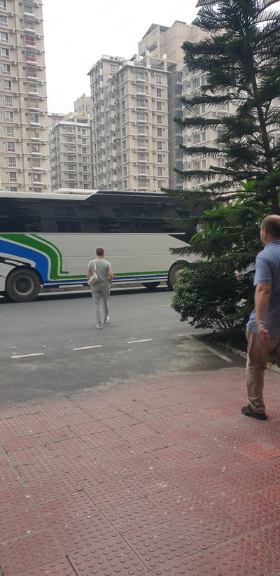 In Chor Sahapur, Bangladesh, a white bus with blue and green stripes is parked on the side of a road. A person in light-colored pants and a grey t-shirt, carrying a white bag, walks away from the bus. Another person, wearing tan pants and a blue t-shirt, stands on a red tiled sidewalk in the foreground, facing away from the camera and towards the bus. Behind the bus, multiple tall, modern apartment buildings with many windows are visible. A large evergreen tree with dense foliage is positioned to the right of the bus, partially obscuring the view of the buildings and the second person. The sky is overcast.