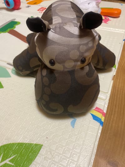 A plush toy, resembling a hippopotamus, lies on a patterned mat. The toy is predominantly brown and beige with a swirling pattern. It has black button eyes and floppy ears. The mat is white with a textured pattern and colorful graphics, including a green leaf shape and a rainbow motif. In the background, out-of-focus, are some children's toys, including a red and blue object and an orange object. The scene is set indoors, with a wooden surface visible to the right of the mat.