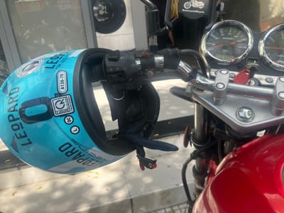 A turquoise "LEOPARD RACING" motorcycle helmet is hanging by its chin strap from the left handlebar of a red Suzuki motorcycle in Ngũ Hành Sơn Ward, Vietnam. The helmet is inverted, displaying "LEOPARD RACING" text, markings "A138-79", "QUATEST 2", "QC", and size "L" stickers. The motorcycle features chrome handlebars, a black left-side control module, and a dashboard with two circular gauges, one visibly labeled "SUZUKI" with a speedometer indicating up to 140 km/h. A key with a red plastic fob is inserted into the ignition. A black phone holder labeled "Motorcycle/Bicycle" is mounted on the handlebars. The motorcycle is parked on a light-colored paved surface, with tree shadows visible. A blurred reflection in the background shows a building interior, including a circular object resembling a washing machine drum with the number "01" visible. No individuals are present.