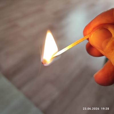 A close-up shot shows a hand holding a lit match, its flame casting a warm glow on the skin. The background is a blurred, neutral surface, possibly a wooden floor or table, with soft lighting. The focus is on the match and the hand, highlighting the texture of the skin and the intensity of the flame. A thin wisp of smoke curls from the matchhead. In the bottom right corner, the date "23.06.2026" and time "19:28" are visible, suggesting the photograph was taken in the evening. The overall mood is intimate and focused, emphasizing the simple act of lighting a match.