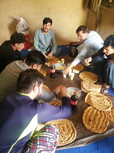 A group of seven men are gathered indoors, seemingly sharing a meal on a large brown sheet laid on the floor. A variety of flatbreads are arranged on the sheet, along with small cups, a bowl containing a white substance, and two bottles of Coca-Cola. Some men are reaching for the food.

The men are wearing a mix of casual clothing, including button-down shirts, t-shirts, and traditional Afghan clothing. The interior appears to be a simple room with unadorned walls. No specific details indicate the location as Arghandab, Afghanistan.
