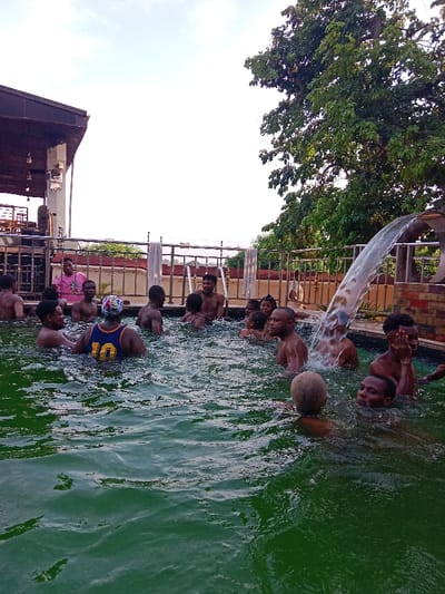 The image captures an outdoor scene of a group of approximately fifteen to twenty people, predominantly young adults and adults, gathered in a swimming pool. The pool water is notably green and shows significant ripples, indicating movement and activity.

Most individuals are submerged in the water up to their chests or shoulders, with many men appearing bare-chested. They are actively interacting, with some looking towards each other in conversation, some gazing towards the camera, and others engaged in various activities within the water. Notable individuals include a person seen from the back wearing a blue and yellow basketball jersey with the number "10" on it, positioned towards the left-center. Another person wears a colorful, patterned swim cap or head covering. On the far right, an individual shields their face with both hands from the splash of a cascading waterfall feature that flows into the pool from a stone or brick structure at the pool's edge. Closer to the foreground on the right, a person is visible with a light-colored, spherical object, possibly a deflated ball or a coconut, floating near their head. The expressions on many faces suggest enjoyment, relaxation, and sociability.

The pool area features a metal railing surrounding its perimeter, and a white towel is draped over this railing towards the left. In the upper left quadrant of the image, a portion of a building or an outdoor structure with a dark roof and supporting pillars is visible. This structure appears to be an adjacent facility, possibly a covered patio or lounge area. The upper right quadrant is dominated by a large, mature tree with abundant green foliage. The sky above is bright and clear, suggesting a daytime setting. Beyond the pool railing and structure, the background appears hazy and less defined, with distant landscape or buildings under the bright sky. No text is visible in the image. The overall impression is one of a cheerful and active social gathering in a recreational swimming environment.