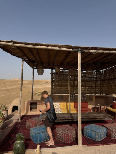 In Agafay, Morocco, a man stands on a large, intricately patterned red rug, holding a mobile phone. He is dressed in a grey t-shirt and brown shorts, with a black bag slung across his body. The rug is adorned with numerous poufs in red and blue, each featuring a distinct geometric pattern.

The scene is set under a rustic, thatched roof supported by wooden poles, providing shade over a seating area. Behind the poufs, a low, dark wooden table sits on the rug. Further back, a built-in bench with cushions in yellow, red, and patterned fabric lines a wall. Decorative woven plates are mounted on the wall above the bench.

The environment is arid, with a desert landscape visible in the background. Hills and sparse vegetation characterize the terrain under a clear blue sky. To the left, a dome-shaped structure, possibly a traditional oven or dwelling, is partially visible. In front of it, potted plants, including an aloe vera, add a touch of green to the dry surroundings. The overall impression is one of a desert-style lounge area.