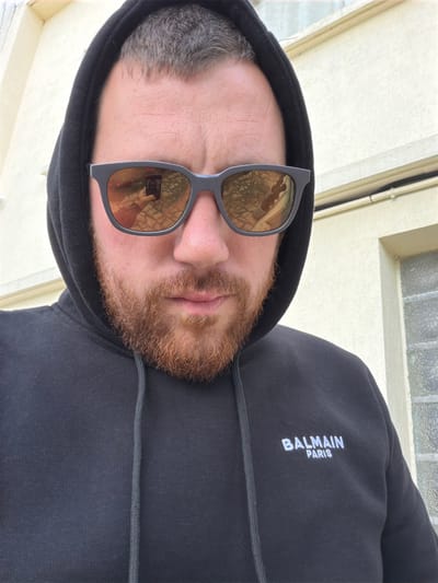 A man wearing sunglasses and a black hooded sweatshirt stands in front of a light-colored building. The sweatshirt has "BALMAIN PARIS" embroidered in white lettering on the left chest. His reflection is visible in the sunglasses, showing him holding a phone. The environment appears to be an urban setting, possibly Bondy, France.