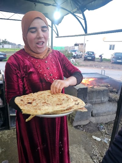 A woman wearing a hijab and maroon dress smiles, holding freshly baked flatbread on a silver plate. In the background, a fire burns in a large outdoor oven constructed of stacked stones. The scene takes place outdoors, possibly near a street with parked cars. The image is slightly rotated.