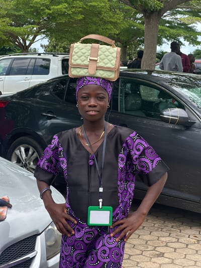 In Itoga, Nigeria, a young girl stands in front of a dark-colored sedan with her hands on her hips. She wears a black and purple patterned traditional outfit and a matching headscarf. A light green quilted handbag rests on top of her head. A green lanyard with an ID badge hangs around her neck. To her left, a silver car is partially visible. In the background, trees and other vehicles, including a white SUV, are present.