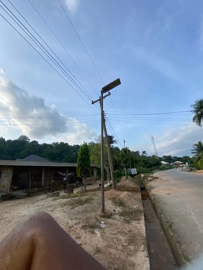 The image depicts a semi-urban street scene in Igarra, Nigeria, under a partly cloudy sky. The foreground shows unpaved, dry, light brown earth with sparse vegetation and minor debris, partially obscured by a blurry, dark brown human limb in the lower left corner.

A narrow, paved asphalt road begins in the mid-ground and extends into the distance, curving slightly right. Parallel to the left edge of the road, a recently excavated, rectangular dirt trench or drainage channel runs lengthwise. Adjacent to this trench, a white sign with blue text, partially reading "ENWAN," is visible.

A wooden utility pole stands prominently in the mid-ground on the left, supporting multiple black electrical wires that traverse the upper portion of the frame. Attached to this pole is a rectangular solar panel street light, angled upwards. To the left of the pole, a tall, slender evergreen tree stands.

Behind the utility pole and tree, on the left side, is a single-story building with a dark, sloped roof and multiple barred openings or windows. Its exterior walls are light brown. Two individuals are present near this building; one person in dark clothing walks away from the structure towards the right, and another is partially visible closer to the building's entrance.

The background features a dense expanse of dark green trees and foliage, forming a hillside behind the structures on the left. On the right side of the road, scattered palm trees and other vegetation obscure additional, smaller buildings. In the far distance, a red and white lattice communication tower stands against the sky, visible above the tree line. On the paved road in the background, two motorcycles are discernible, with individuals near them.