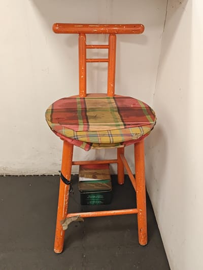An orange wooden stool with a plaid seat is positioned against a white wall. A box is placed on the floor beneath the stool, supported by some of its legs. The stool has a worn appearance, with chipped paint and frayed fabric on the seat. The wall behind the stool is textured and has some faint markings. The floor is a dark, textured surface. The overall scene suggests a slightly aged and utilitarian setting, possibly in an interior space within the city of Settimo Torinese, Italy.