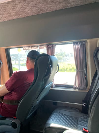 The image shows the interior of a vehicle, likely a bus or van, with a person sitting in the foreground. The person is wearing a dark baseball cap and a maroon t-shirt, and is leaning forward, with their head obscured by a large, black case that appears to be for a musical instrument, possibly a cello or bass. The case is resting on the person's lap and is angled towards the viewer.
The setting is a vehicle interior with upholstered seats and windows. To the left, there is a seat with a diamond-stitched pattern. To the right, a quilted grey seat is visible. The windows have dark pleated blinds. Through the window in the middle ground, a blurry view of trees and possibly buildings can be seen, suggesting it's daytime and the vehicle is in motion. Sunlight is coming through the window, illuminating the scene.
On the window in the foreground, there is text in white lettering on a yellow background, which reads "ЗАПАСНОЙ ВЫХОД" (Emergency Exit). Below this, there is a smaller yellow diamond shape with some text that is difficult to read. The overall impression is one of travel. The emotions conveyed are neutral, focusing on the act of carrying the instrument case during transit. The time of day appears to be daytime, and the weather is likely sunny, given the bright light. The location context of Białystok, Poland is provided as a hint, but there are no specific visual cues within the image that confirm this location.