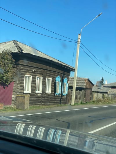 The image depicts a street scene in Kuytun, Russia, on a clear, sunny day. In the foreground, the reflection of the environment is visible on the dark surface of a vehicle's dashboard or hood, showing inverted images of windows and parts of the street.
The primary subject is a single-story dark brown wooden log house with a grey corrugated metal roof, positioned on the left side of an asphalt road. The house features three vertical white-framed windows on its left side and two vertical windows with ornate, light blue wooden shutters on its right side. A house number "42" is visible to the left of the leftmost white window. To the far left, a section of a deep red corrugated metal fence or gate is visible, with green shrubbery partially obscuring it. A rectangular, grey concrete-like structure rests on the ground below the leftmost windows.
Adjacent to the house, a light grey utility pole stands upright, supporting multiple black electrical wires that span across the upper portion of the frame. A modern streetlight fixture extends diagonally from the pole to the right. Beyond the utility pole and a wooden fence, additional residential or utilitarian structures with grey corrugated metal roofs are visible. In the distance, a white chimney on a building emits a plume of light grey smoke, and a small, conical green tree stands nearby. The sky is clear and bright blue, without any visible clouds. No individuals or animals are present in the scene. The asphalt road in the foreground features a distinct solid white line marking the edge of the visible lane.