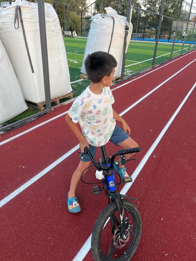 A young boy sits on a bicycle on a red running track with white lane markings. He is wearing a white t-shirt with a colorful graphic print and blue shorts. He has on blue clogs with Mickey Mouse on them. The boy is facing to his right, looking away from the camera. In the background, there are large white bags stacked, possibly for construction or storage, and a fenced-in sports field with green turf and blue courts. The location is identified as Koroviia, Ukraine.