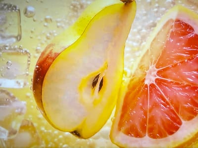 The image shows a close-up, macro shot of a sliced pear and a slice of grapefruit submerged in a bubbly, yellow liquid, possibly a drink or soda. Ice cubes are visible in the background on the left side of the image. The pear is cut lengthwise, revealing its seeds. The grapefruit slice showcases its segmented flesh and juicy texture. The lighting highlights the water droplets and bubbles clinging to the fruit and ice. There are no people or specific landmarks present in the image. The scene is aquatic and food-focused. There is no information to suggest the location in the city of Shakhovskaya, Russia.