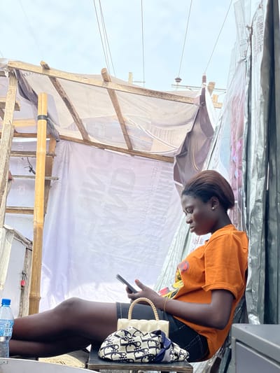 The image depicts a young woman in an orange t-shirt and black shorts standing under an awning constructed from wood and white fabric. She is looking down at her phone, which she holds in her right hand. A white handbag with a patterned design is hanging from her left arm. A bottle of water is visible on a surface below her.
The scene appears to be outdoors, possibly in a market or street stall, given the makeshift shelter and the presence of utility lines in the background. The lighting suggests it is daytime, though the sky is overcast, hinting at cloudy weather. The setting seems to be in Ojomu, Nigeria, as indicated by the location context. There is some visible text on the white fabric of the awning, but it is largely obscured and difficult to read. The woman's posture and focus on her phone suggest she is engaged in a personal activity, possibly communication or browsing.