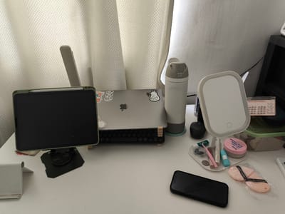 The image depicts a white desk with various personal items arranged on its surface. A tablet, held by a green case and stand, is positioned on the left side of the desk. To its right, a silver laptop is open, displaying a keyboard with round keys. A white and gray water bottle stands behind the laptop. In the center of the desk, a makeup mirror with a light ring is present, surrounded by small cosmetic items including a pink powder compact, makeup brushes, and a razor. A black smartphone lies on the lower right side of the desk. The background consists of a textured white curtain and a white wall. The overall impression is of a tidy workspace, possibly a personal vanity or study area. The location provided, General Trias, Philippines, suggests a domestic or residential setting.