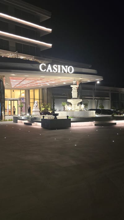 The image shows the exterior of a casino at night in Agios Epiktitos, Northern Cyprus. The building has a modern facade with illuminated horizontal lines on its upper levels. A large, illuminated sign spelling "CASINO" is prominently displayed above the entrance. The entrance area features a wide, covered walkway with a grid-like ceiling illuminated from within. In front of the entrance, there is a large, multi-tiered white fountain with decorative sculptures, including lions at its base and a central figure. To the left of the fountain, there is a white statue of a seated lion. Landscaping with green bushes and small trees surrounds the fountain and the statues. A person dressed in a dark suit and tie stands near the entrance, facing the camera, acting as a security guard or doorman. Another person is partially visible through the glass doors of the entrance. The ground in front of the casino is paved with large, dark tiles, with small circular lights embedded in the surface. Several dark-colored vehicles are parked to the right of the fountain. The overall atmosphere is one of a well-lit, upscale establishment at night.