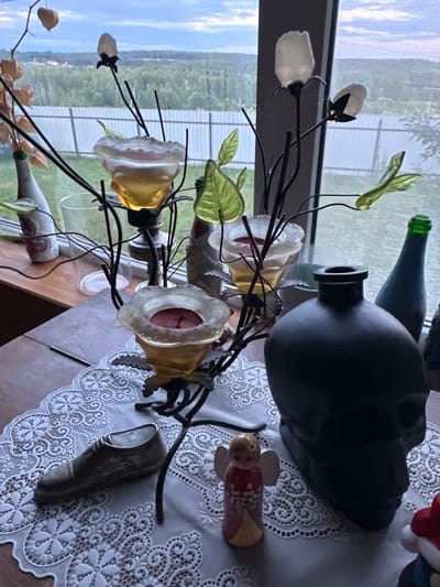 A still life composition is arranged on a wooden table, partially covered by a white lace doily. The arrangement includes a decorative metal candelabra holding three glass candle holders, one with a red candle. To the left of the candelabra is a small, metallic shoe figurine. To the right of the candelabra, a dark, skull-shaped vase dominates the foreground. Between the shoe and the skull vase sits a small, ceramic angel figurine. In the background, through a window, a rural landscape with trees and a fence is visible under a cloudy sky. The scene is indoors, with the window providing a view of the exterior. There are no people or animals present in the image.