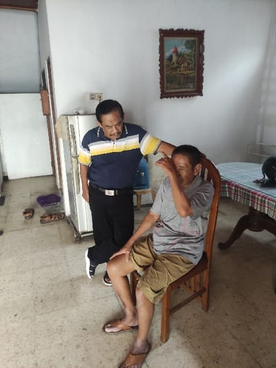 This media file captures an indoor scene, likely a living room or common area in Lamongan, Indonesia. The image is oriented vertically but depicts two men in what would naturally be a horizontal setting.
On the left, a man with dark skin and short dark hair stands, wearing a dark blue polo shirt with yellow and white stripes, black trousers, and black and white sandals. He has a watch on his left wrist and a silver belt buckle visible. His gaze appears directed towards the right side of the frame.
On the right, another man, also with dark skin and short dark hair, is in a relaxed or resting position, leaning against a wooden chair. He wears a grey t-shirt, brown shorts, and brown sandals. His right arm is bent, with his hand near his face or head, suggesting he might be rubbing his eye, resting, or in a pensive state.
The room features light-colored, possibly concrete or tiled, flooring with some visible marks. The walls are white. A dark wooden framed painting depicting a landscape with a building hangs on the wall behind the standing man. Below him, there's a white electrical outlet, a light fixture, and what appears to be a large white appliance, possibly a refrigerator. Part of a table with a plaid tablecloth and a black helmet or bag is visible in the upper right background. A blue container and a purple fabric item are also seen on the floor.
The scene suggests a casual daytime gathering or moment of repose. There is no visible text in the image. Emotions are not overtly clear, with the standing man appearing neutral and the leaning man's expression obscured.