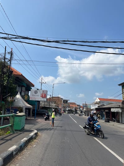 This is a vertical, eye-level, medium shot of a street in Kludan, Indonesia, on a bright, sunny day. The street is paved and has a white dividing line. On the left side of the street, there is a sidewalk with a green railing and a green trash bin. Beyond the sidewalk, there are buildings and trees. One building has a white tent in front of it and a large advertisement banner. Overhead, there are many electrical wires. On the right side of the street, there are more buildings and storefronts. In the middle of the street, a person wearing a yellow vest and a hat is standing and holding a yellow and red stick. Further down the street, a person is riding a blue motorcycle with a black accent. The sky is a clear blue with a few scattered white clouds.
