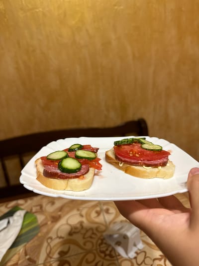 Two open-faced sandwiches are presented on a white plate. Each sandwich consists of a slice of bread topped with mayonnaise, a slice of sausage, a slice of tomato, and slices of cucumber. The background consists of a textured golden wall and a portion of a wooden chair. The plate is held by a person's hand. The setting appears to be indoors, potentially a home, and the details do not provide any specific information about the city of Chemerne, Ukraine.