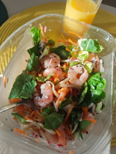 The image shows a close-up, overhead view of a shrimp salad in a clear plastic takeout container, placed on a yellow placemat. A glass of orange juice is visible in the upper right corner of the frame. The salad contains cooked shrimp, shredded carrots, leafy greens, and possibly radish and other finely chopped vegetables, garnished with black sesame seeds. A fork is partially visible on the left side of the container, resting on the salad. The lighting suggests it is daytime, possibly a meal being consumed outdoors or near a window, with sunlight casting shadows. There are no people or animals in the image. The setting appears to be a casual dining environment. The image was not taken in Tilburg, Netherlands.