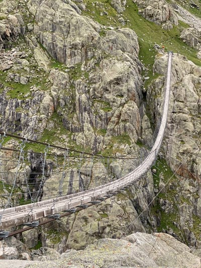 The image presents the Trift Bridge, a pedestrian suspension bridge, traversing a rugged, rocky gorge near Innertkirchen, Switzerland. The bridge features a walkway constructed from wooden planks, supported by multiple steel cables, and flanked by wire mesh safety barriers. It extends diagonally upwards from the lower right foreground towards the upper left, anchored into the steep mountain face. The surrounding environment consists of prominent grey and brown rock formations and boulders, interspersed with patches of vibrant green grass and moss. At the far end of the bridge, two small human figures are discernible; one appears to be standing, and the other is positioned as if resting or sitting. Adjacent to the bridge's far anchor point, on a grassy slope, a distinct orange spherical object is visible. The immediate foreground displays rough, lichen-covered rocks, establishing the perspective from the bridge's near approach.