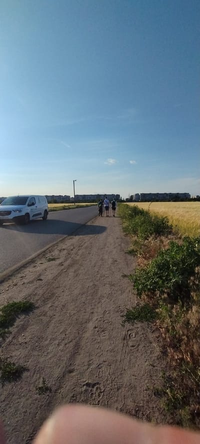 The image depicts a street scene with a clear blue sky and a field of golden crops. In the foreground, a dirt path runs parallel to an asphalt road. A white van is driving on the road, and three individuals are walking away from the camera on the dirt path. Behind them, a line of apartment buildings stretches across the horizon, suggesting an urban or suburban environment. The overall impression is a peaceful, daytime scene, possibly in the city of Sokolivske, Ukraine, given the context.