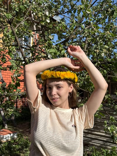 A young woman with light brown hair and a yellow dandelion crown stands amongst the branches of a blossoming tree. Her arms are raised, framing her face. She wears a striped t-shirt and a gold hoop earring is visible. The background includes green foliage, patches of blue sky, wooden fencing, and a red brick building. The location is Кемуль, Russia.