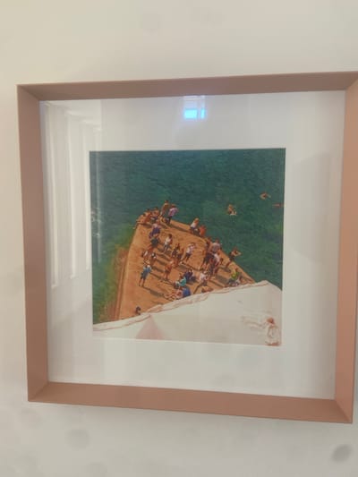 A framed photograph depicts a group of people gathered on a sun-drenched, rocky outcrop overlooking a body of turquoise water. The perspective is from above, looking down at the scene. The people are dressed in summer attire, suggesting warm weather and a leisure activity. Some individuals are standing, observing the water, while others appear to be preparing to enter it. One person is captured mid-jump or dive into the water, with a splash visible below. The water itself is clear, with visible ripples and a vibrant green hue, indicative of shallow depths and sunlight. The rocky surface is a warm, sandy color, contrasting with the cool tones of the water.
On the right side of the image, a section of white fabric or drapery is visible, creating a surreal and possibly symbolic element within the otherwise realistic scene. This fabric appears soft and flowing, adding a different textural quality. The overall impression is one of a lively summer day at a coastal or lakeside location, filled with anticipation and the joy of outdoor recreation. The time of day appears to be midday or early afternoon, given the strong sunlight casting shadows on the rocks. There is no visible text within the photograph itself. The location context suggests this could be a popular spot for swimming or cliff diving in Matosinhos, Portugal.