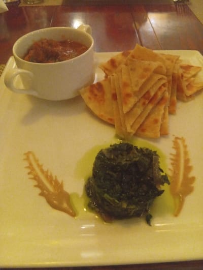 A square plate holds a culinary arrangement: a white mug filled with a tomato-based stew sits beside a stack of triangular flatbreads. Below, a mound of dark green, possibly sautéed, spinach glistens with oil, flanked by two ornate sauce designs resembling feathered fronds. The presentation is artistic, suggesting a fine dining experience. The color palette is warm, with earth tones dominating the visual.