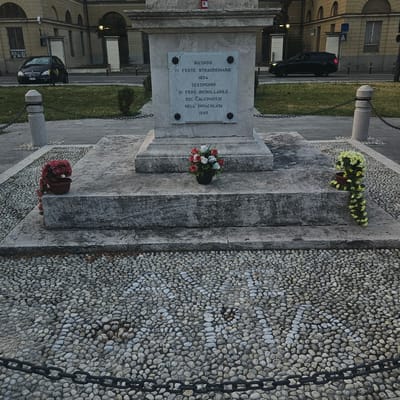 The image shows a monument with a plaque inscription. The inscription reads: "RICORDO DI FESTE STRAORDINARIE 1854 TESTIMONIO DI FEDE INCROLLABILE DEI CALCINESI NELL'IMMACOLATA 1895." This translates to: "REMINDER OF EXTRAORDINARY FESTIVITIES 1854 TESTIMONY OF UNWAVERING FAITH OF THE PEOPLE OF CALCINATE IN THE IMMACULATE 1895." The monument is made of stone and is located outdoors, on a base covered in small, smooth pebbles. There are floral arrangements placed at the base of the monument. In the background, there are buildings and cars, suggesting an urban or town setting, likely in Calcinate, Italy, given the inscription. The scene appears to be a public space, possibly a square or a park. The lighting suggests it might be dusk or an overcast day. There are no people visible in the frame.