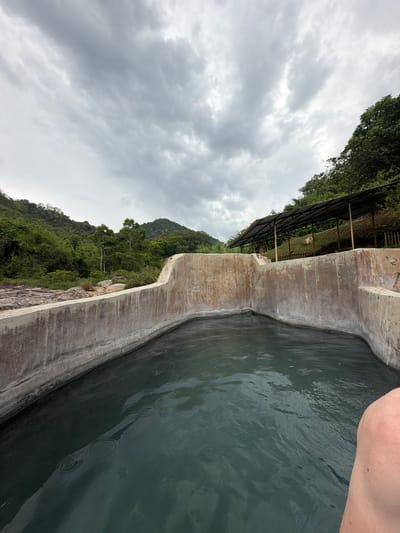 The image shows a man's knee and leg in the lower right corner, partially submerged in a dark, murky pool of water. The pool is contained within a concrete structure with rough, weathered walls. In the background, a lush, green hillside is visible, with a dense forest covering its slopes. A dark, open-sided shelter with a sloped roof stands on the hillside above the pool. The sky is overcast with heavy, grey clouds. The scene appears to be an outdoor hot spring or thermal pool located in the natural landscape of Khanh Vinh, Vietnam, given the tropical foliage and mountainous terrain.