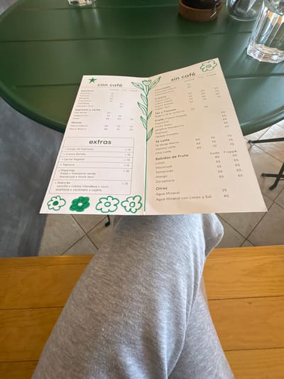The image shows a menu with a green floral design, listing coffee, non-coffee drinks, and extras. The menu is in Spanish and includes items like "Espresso", "Latte", "Chocolate Clásico", and "Bebidas de Fruta". Prices are listed next to each item. The menu is resting on a green, possibly metal, table or surface. Part of a person's leg, clad in grey sweatpants, is visible in the upper right corner of the image. The menu is from a cafe in Oaxaca City, Mexico.