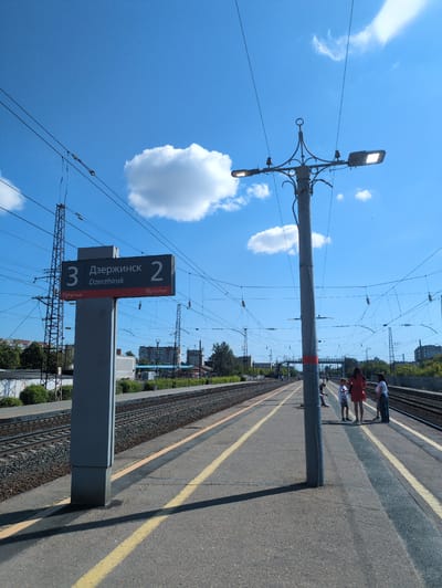 The image depicts a train station platform in Dzerzhinsk, Russia, under a clear blue sky with scattered white clouds. A tall, gray pole with multiple lights and electrical insulators stands prominently on the platform. To the left, a directional sign displays "3 Дзержинск Dzerzhinsk 2" and "Путь track" in both Russian and English. The platform is paved with asphalt and marked with yellow lines. Railroad tracks run parallel to the platform on both sides. In the background, buildings and trees are visible, along with overhead power lines and electrical pylons. Three people are standing on the platform near the central pole: a woman in a red dress and two children. Another person is seated on the platform in the middle ground, to the left of the pole. The scene appears to be a sunny day at a functional, outdoor transportation hub.