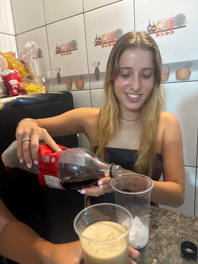 A young woman with long, light-brown hair smiles as she prepares a drink. She holds a large bottle of dark soda in one hand, ready to pour. To her right, two clear plastic cups are visible: one filled with ice and the other containing a light-colored, frothy beverage. The scene appears to be indoors, in a kitchen with decorative tile walls. Other plastic cups and snacks are visible in the bottom left corner.