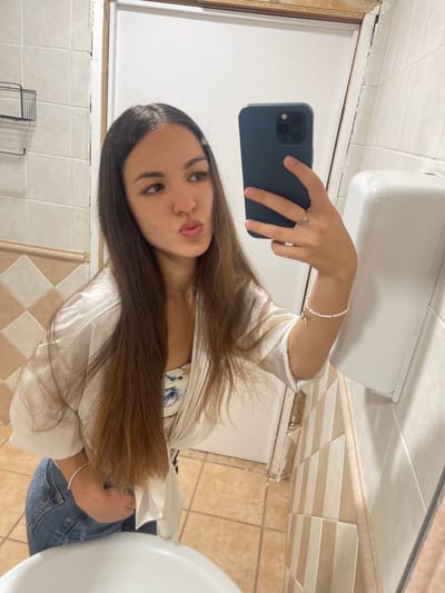 A young woman is taking a selfie in a bathroom. She is holding a dark-colored smartphone in her right hand. She has long brown hair and is wearing a white, long-sleeved shirt with a hint of a floral-patterned top underneath, and blue jeans. She is also wearing a ring and a bracelet. The bathroom has beige and white tiled walls, a white sink and a white hand dryer. There is a door in the background. The location is specified as Paiporta, Spain.