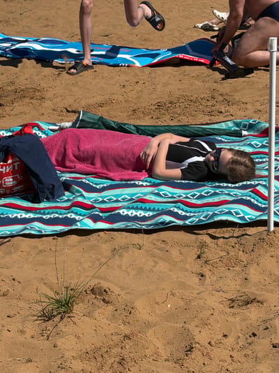 A person lies on a beach blanket on the sandy shore of Malakhovka, Russia, with a pink towel draped over their lower body. The person, wearing sunglasses and a black and white shirt, has their arms crossed. Other beach items, including a blue bag and a deflated inflatable, are visible in the background. The image captures a sunny day at the beach.