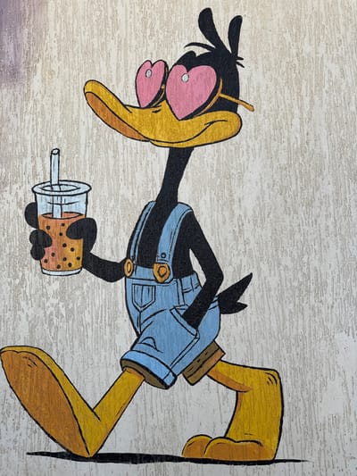 A cartoon character resembling Daffy Duck is depicted in a stylized, hand-painted artwork. The character is primarily black with a long, yellow-orange bill and large, flat feet. It is dressed in light blue overalls, complete with golden buttons and straps. Its eyes are noticeably pinkish-red, conveying a sense of extreme tiredness or a deliberate artistic choice. A thin, yellow, straw-like object protrudes from behind the character's head.

In its left hand, the character holds a clear plastic cup containing a brown beverage with several dark, circular boba pearls and a white straw. The character appears to be in a relaxed or lounging position, with its body extending horizontally across the frame.

The artwork is painted directly onto a textured, light beige or off-white surface, likely a wall or a similar architectural element, with the texture of the underlying surface visible through the paint. The overall aesthetic is cartoonish and expressive. There is no visible text in the image. The scene is situated in Safaga, Egypt.