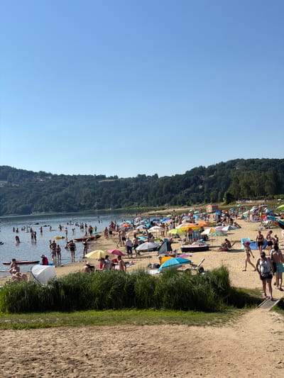 This media file depicts a vibrant summer day at Gródek nad Dunajcem, Poland. The scene is a bustling beach packed with numerous people enjoying the water and the sand. A clear blue sky suggests fair weather, likely mid-day given the bright sunlight and shadows.

The subjects are primarily people, many of whom are sunbathing on the sandy shore or wading and swimming in the lake. Colorful umbrellas dot the beach, providing shade for some of the beachgoers. Objects visible include beach chairs, towels, and a few surfboards or paddleboards in the water. The setting is a lakeside beach bordered by a dense, green forest that rises up a hillside. A sandy path runs alongside the grassy edge of the beach, leading further down the shoreline.

The activity is clearly recreational, focused on enjoying a warm day at the beach. People are engaged in various activities: lounging, swimming, walking along the shore, and interacting with each other. The overall mood appears relaxed and lively, typical of a popular summer vacation spot. There are no visible text elements in the image.