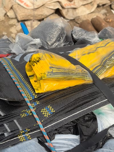 The scene, located in Jakande, Nigeria, depicts a detailed close-up of an accumulation of various materials, indicative of a storage or informal commercial setting. In the foreground and midground, a substantial stack of folded black woven polypropylene bags is prominent. Each bag displays a repeating horizontal band pattern featuring small blue and yellow checkered squares. A multi-colored woven rope, composed of interlaced blue, orange, and white strands, lies diagonally across the stack of black bags. Positioned on these black bags is a clear plastic bag enclosing a tightly packed stack of bright yellow plastic bags, each folded flat. A black strap or band runs horizontally across the yellow bags. In the midground and background, multiple translucent plastic bags filled with dark, lumpy contents are visible. Behind these, an irregular pile of light-colored, likely white or off-white, woven sacks forms a backdrop; some sacks display partial text, such as "FLOUR," and appear to contain solid material. To the far right background, several dark, cylindrical objects, consistent with cut wood logs or pipes, are partially discernible. A blue cylindrical object is also visible in the upper left background. No individuals are present, and no active interactions or actions are depicted. The environment suggests an outdoor or semi-enclosed area characterized by a mixture of textures and industrial or commercial materials.