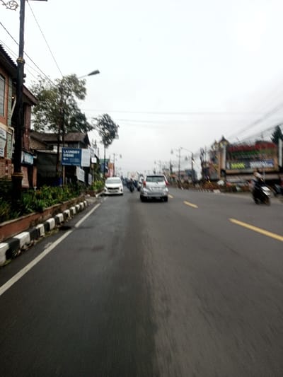 This image depicts an urban street scene in Chagzam_Pam, Bhutan, under an overcast, bright grey sky. A multi-lane asphalt road, appearing damp in sections, extends from the foreground into the distance. On the left, a low concrete curb with black and white painted segments separates the road from a raised planter containing green foliage. A decorative black and gold street pole with overhead utility lines stands on the far left. Behind the planter, multi-story buildings line the street; one structure displays a blue sign with white text, "LAUNDRY DISINI," and smaller, illegible text below.
Vehicular traffic moves away from the viewer on the roadway. A white car is positioned near the left curb, and a silver SUV occupies the middle lane. Further ahead, several motorcycles and other vehicles are present. On the far right, a dark motorcycle with a rider is visible in motion, indicated by blur. The road surface features a white solid line along the left curb and double yellow lines in the middle to right section. In the background, additional buildings with various commercial signs and multiple street lights on poles line both sides of the road, beneath a network of utility wires.