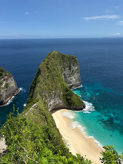 The image depicts an aerial view of Kelingking Beach, located in Bunga Mekar, Nusa Penida, Indonesia. A large, T-shaped headland covered in dense green vegetation extends into the turquoise and deep blue ocean. The headland features a steep, rugged cliff face with visible rock strata. A sandy beach, edged by white foamy waves, nestles at the base of the headland. A walking path is visible along the ridge leading to the beach, and there are 2 people visible in the sand. The sky is clear blue with some wispy clouds.