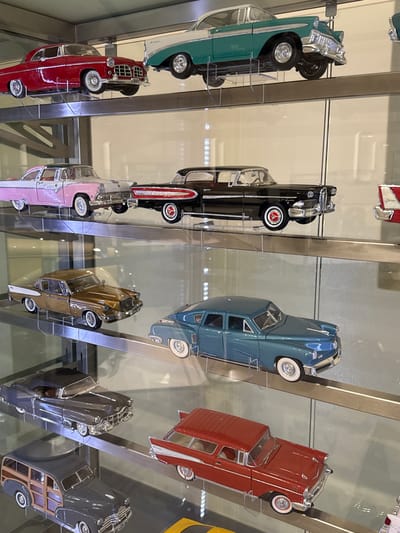 The image shows a display case containing several classic car model replicas. These models are showcased in a vertical arrangement, with each car mounted on a thin metal stand. The cars are predominantly from the 1950s and 1960s, with colors including turquoise, black, red, blue, pink, gold, dark gray, and silver. The display case appears to be made of glass and metal. The background is blurry, suggesting the surrounding environment is out of focus. There are no people visible in the image. There is not enough information to make out the specific location in the city of Stellenbosch Local Municipality, South Africa.
