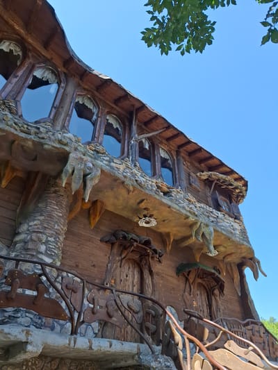 This image depicts a uniquely designed, multi-level building in Lukovit, Bulgaria, characterized by an organic, rustic architectural style. The structure features a prominent, curved wooden roof with visible rafters and multiple irregularly shaped, possibly round or oval, windows on the upper level, behind which white ruffled curtains are visible. Below the upper windows, a textured stone or concrete overhang juts out, adorned with sculpted, root-like or stalactite-like formations that appear to drip downwards.
The building's facade primarily consists of horizontal wooden planks, interspersed with textured stone and concrete elements, including a visible stone-clad column on the left supporting an overhang. Two arched, carved wooden doors, integrated into the organic design, are visible on the lower level, each topped with a small, moss-covered canopy-like feature. A distinctive railing, crafted from curved wood or metal resembling branches and incorporating abstract flat wooden shapes, fronts a lower balcony or terrace. The scene is illuminated by bright sunlight under a clear, intense blue sky, with a cluster of green tree leaves visible in the upper right corner.
