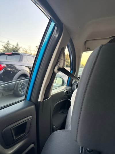 The image provides an interior view from the front passenger side of a vehicle, looking towards the front and outward. The vehicle's B-pillar displays a bright blue exterior color at the window frame, bordered by a black rubber seal and beige interior trim. A beige seatbelt height adjuster is visible on the B-pillar, from which a black seatbelt strap extends downward. The interior of the front passenger door is black textured plastic, featuring a recessed handle. The dark grey or black fabric headrest of the front passenger seat occupies the foreground on the right, with a person's upper body, wearing a light-colored garment, partially visible behind it.
Through the window, a dark-colored SUV or crossover, possibly a Mini Countryman given the distinctive wheel design and body contours, is visible outside. This vehicle displays a red rear taillight and is either stationary or moving slowly in an adjacent lane or behind the observer's vehicle. The exterior environment includes distant sparse trees and a pale sky with a warm, golden gradient near the horizon, consistent with either early morning or late afternoon conditions. The scene depicts a roadway environment within West Windsor, United States; however, specific landmarks or a precise street location are not discernible from the image.