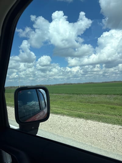 The image shows a view from inside a car. The scene is oriented sideways. A blue sky with fluffy white clouds dominates the view through the car window. A side mirror is visible, reflecting the sky. Outside, green fields extend along the roadside, adjacent to a gravel road. A power pole is seen in the distance. The exact location in Пожарское сельское поселение, Ukraine is indeterminable without further information.