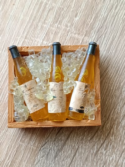 A miniature wooden crate containing three miniature amber-colored bottles filled with a yellow liquid is depicted. The bottles are surrounded by clear, translucent resin pieces shaped like ice cubes. Each bottle has a label with text that is partially obscured by the "ice." The crate and its contents are placed on a light-colored wooden surface with a wood grain pattern. The image is taken from a top-down perspective. There are no people or animals present. The location in the city of Tebras, Latvia is not discernible from the image.