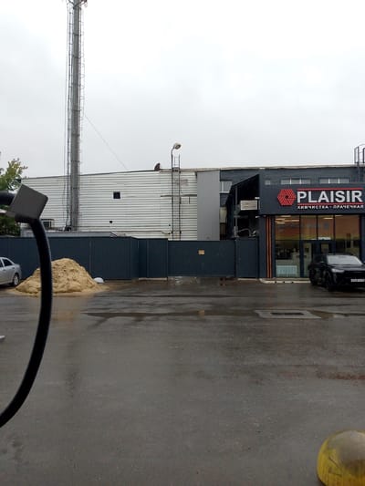 The image shows an outdoor scene under an overcast sky. A large industrial building with a corrugated metal facade dominates the background. A tall, narrow tower with a ladder structure is attached to the building on the left. To the right of the building, a modern storefront with a prominent "PLAISIR" logo and the Russian words "ХИМЧИСТКА - ПРАЧЕЧНАЯ" (Dry Cleaning - Laundry) is visible. A dark-colored SUV is parked in front of the storefront, and a silver car is parked to the left, near a large pile of sand. A black industrial hose or tube extends into the frame from the left foreground. The ground is wet asphalt, suggesting recent rain. The environment appears to be an industrial or commercial area in Saransk, Russia.
