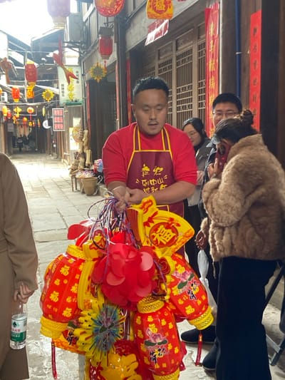 A man wearing a red shirt and a brown apron is holding a bunch of colorful lanterns and paper decorations. He appears to be a vendor in a traditional Asian market or street. Other people are visible in the background, including a woman in a brown coat. Red banners hang overhead. The scene is a busy outdoor setting with a variety of shops and stalls lining a narrow street.